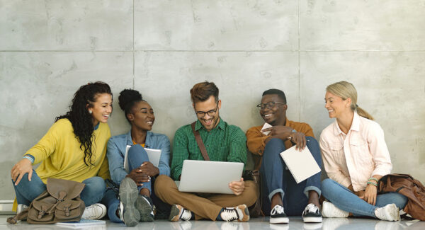Group of happy students surfing the net on laptop while sitting on the floor at university hallway. Copy space.